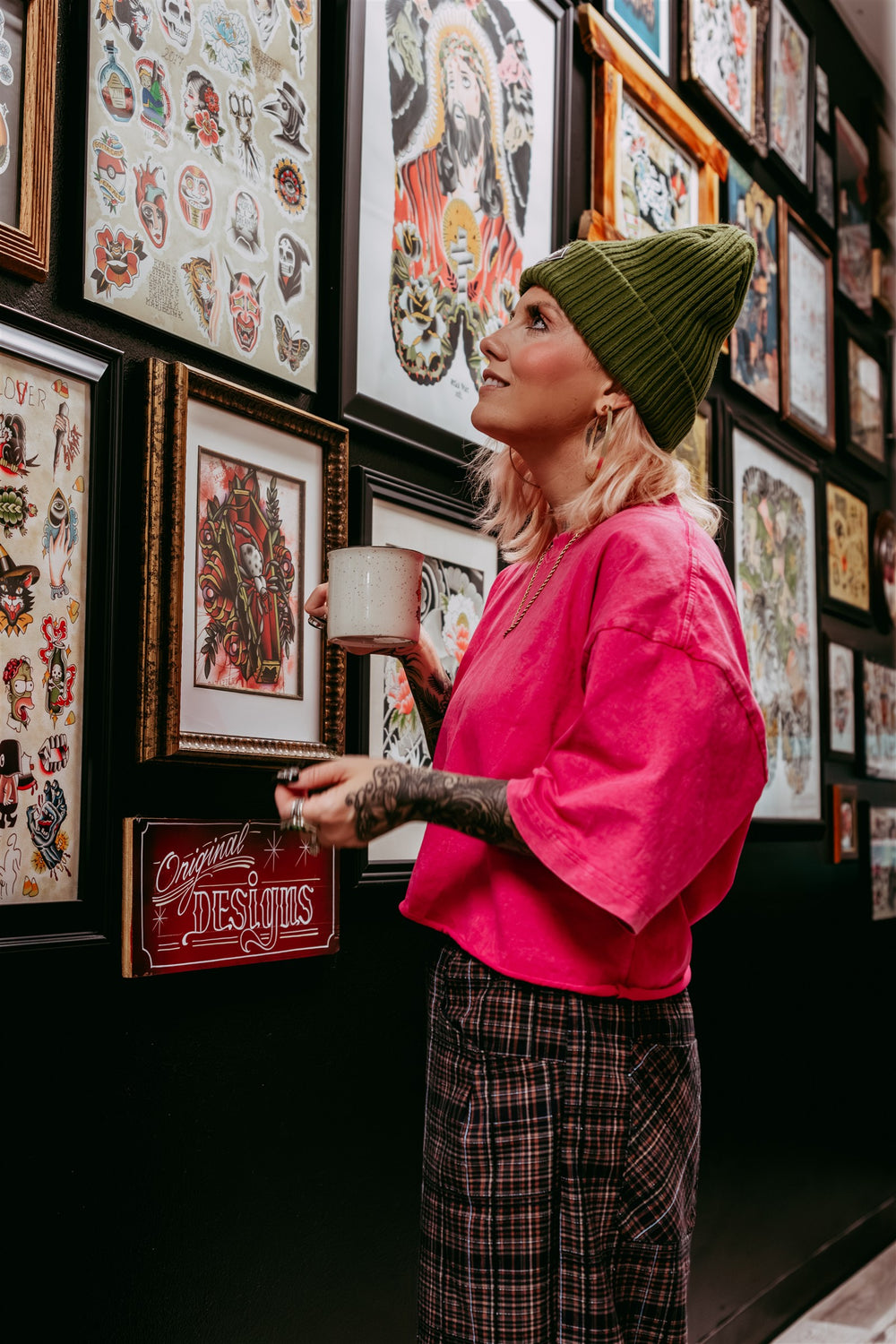 Maribeth holding a mug beside a wall of framed tattoo flash, wearing a bright pink shirt.