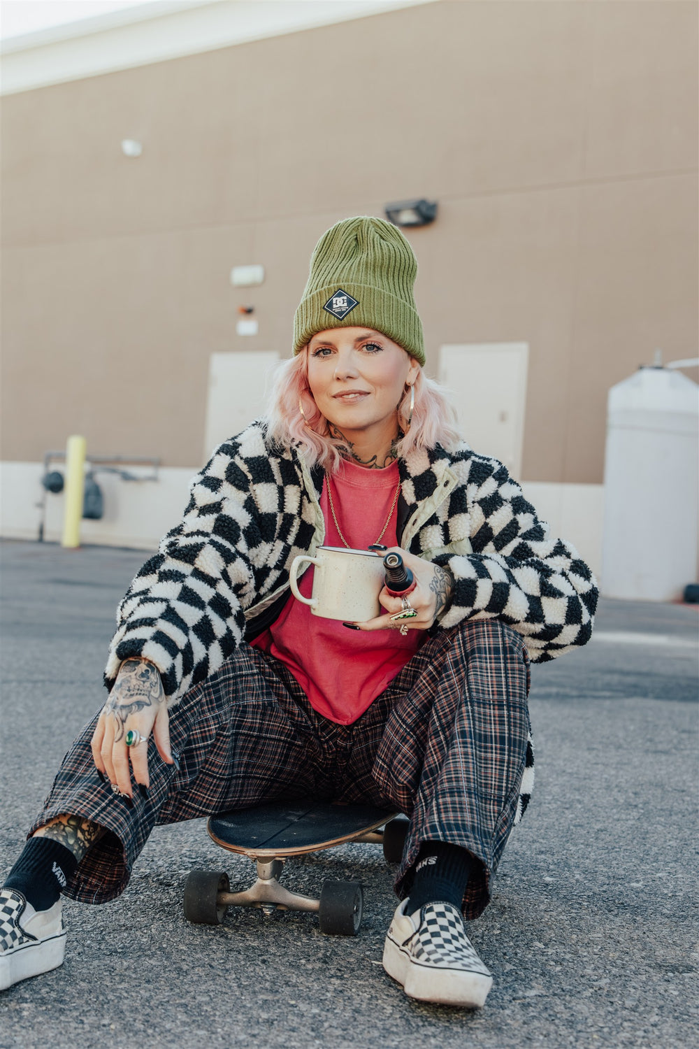 Maribeth sitting on a skateboard with a coffee mug, wearing a checkerboard jacket and green beanie.