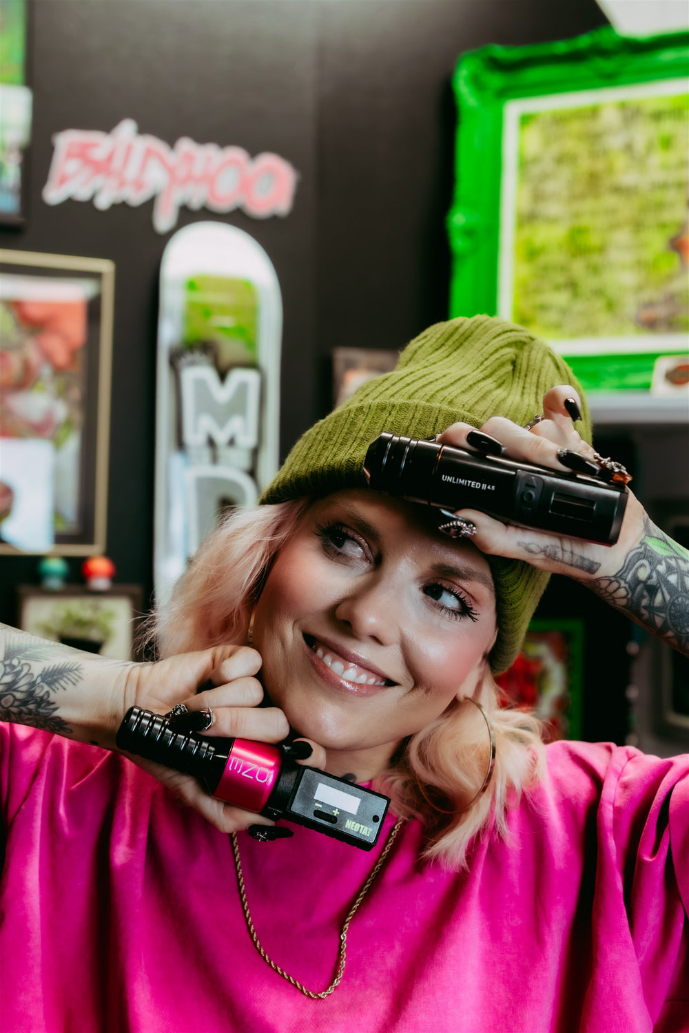 Maribeth smiling in her tattoo studio, holding tattoo machines and wearing a pink shirt and green beanie.